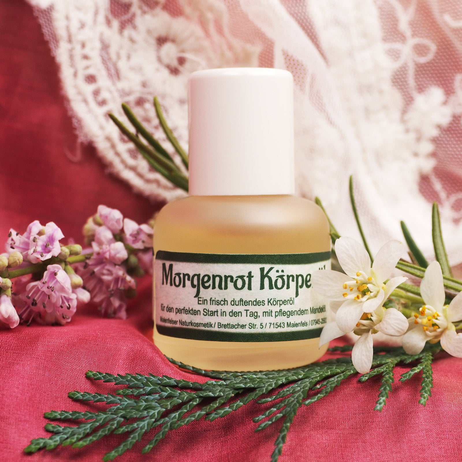 Morgenrot Körperöl in 15ml Glasflasche abgefüllt. HIntergrund weiße Tischdecke mit Spitze , einzelner Tannenzweig sowie rosa und weiße Blüten